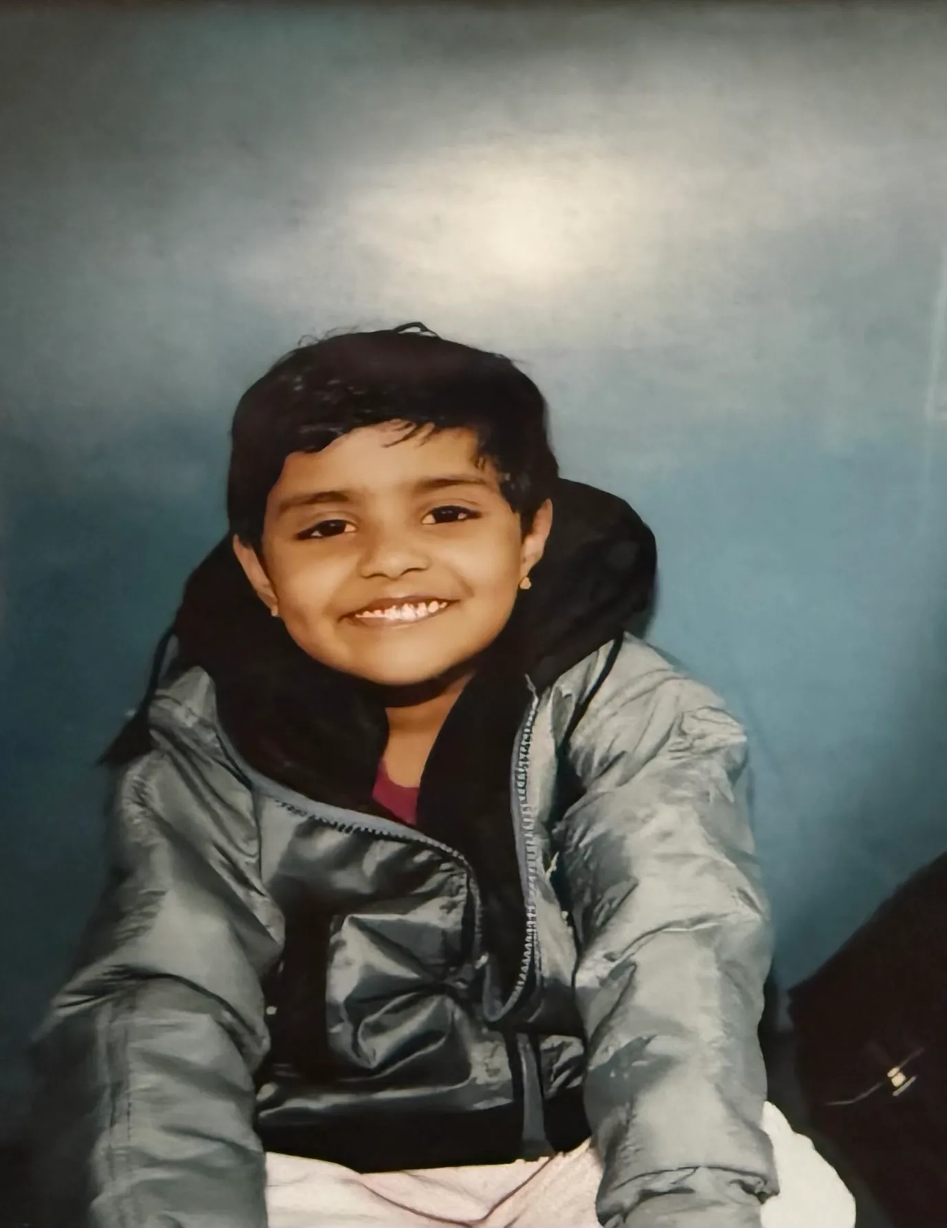 Keren as a child, smiling in a grey puffer jacket against a blue backdrop.