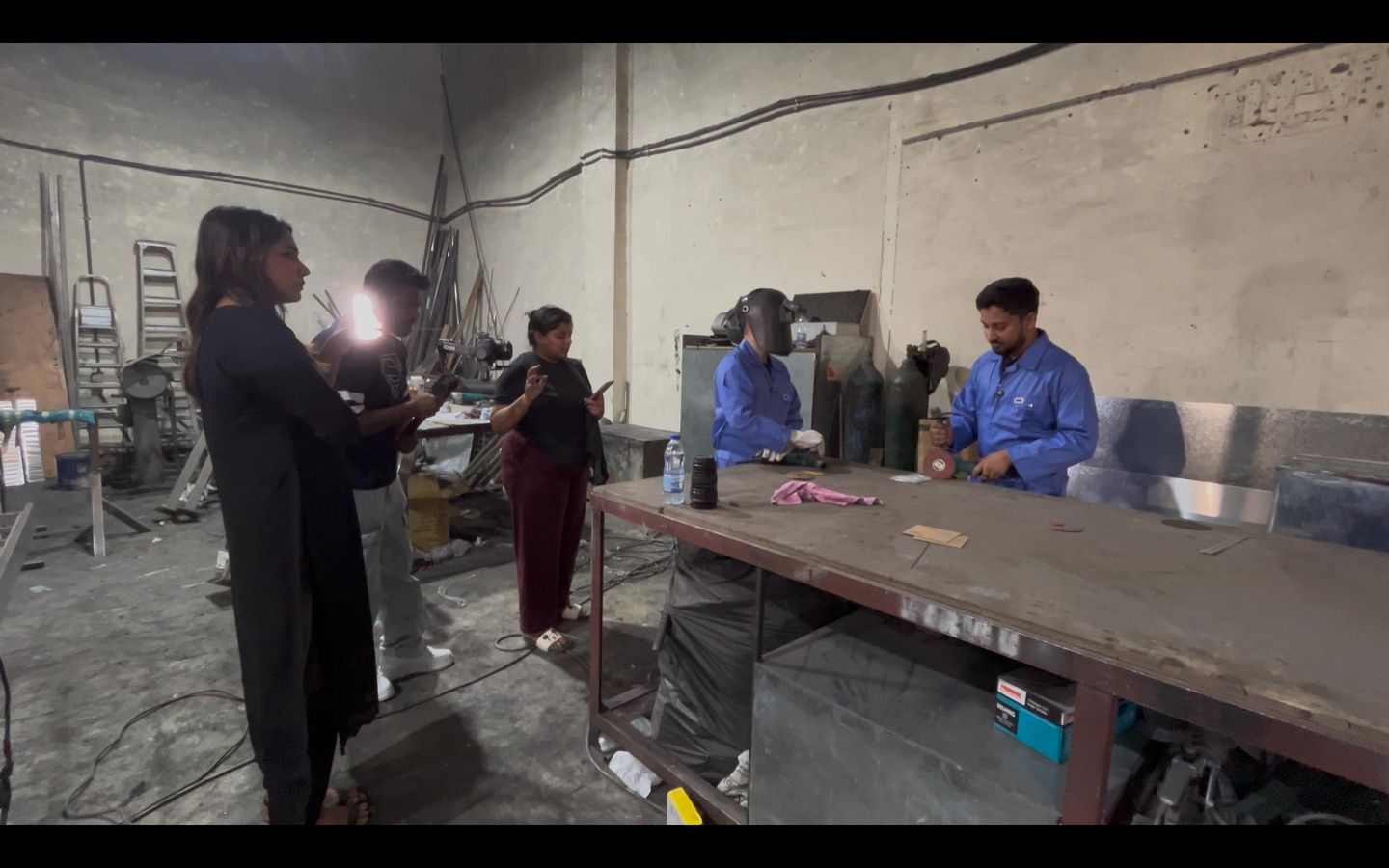 Filming inside a metal workshop: a crew member with a light and camera stands beside a long workbench where two workers in overalls are working.