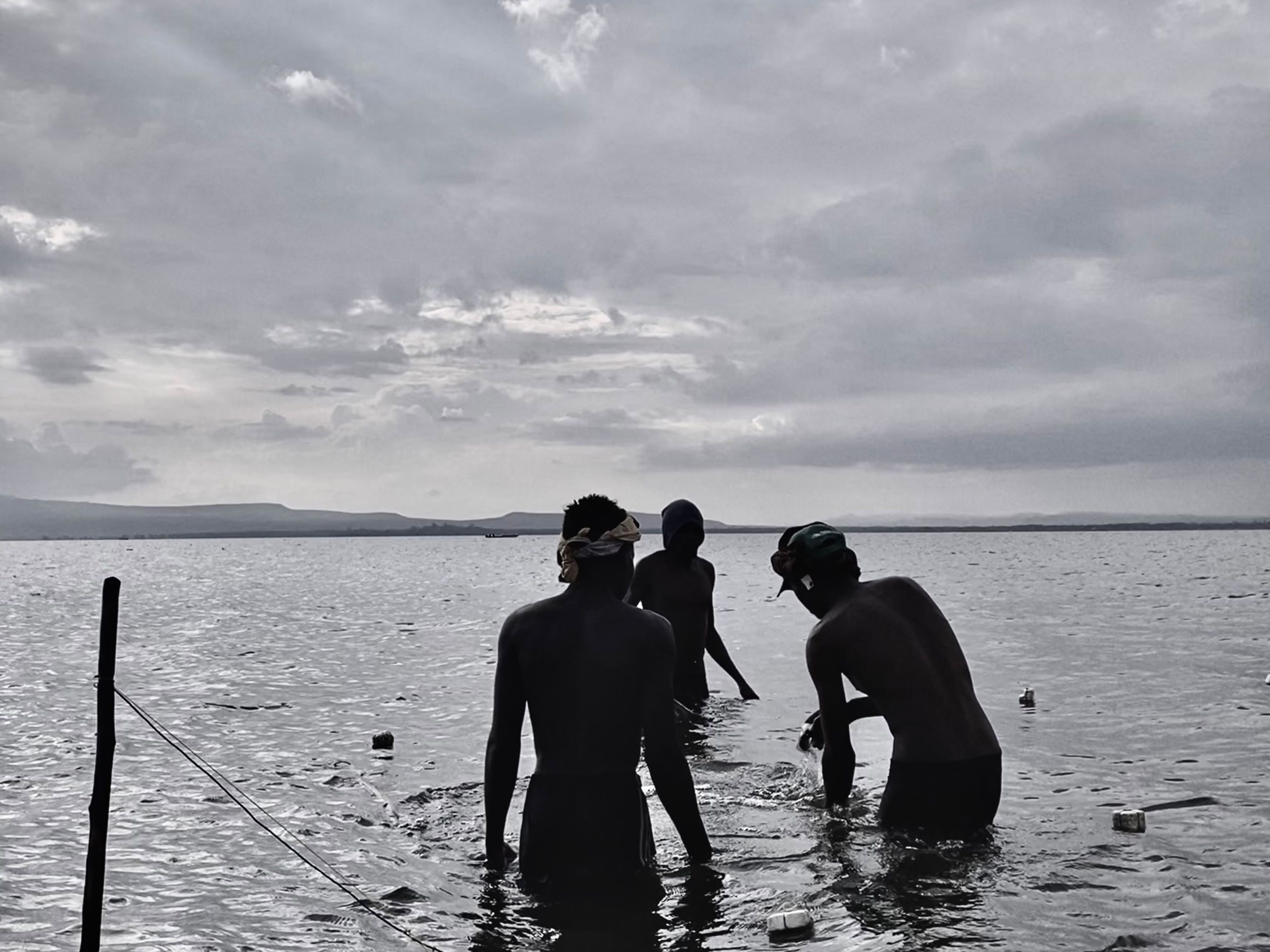 Black and white photo of three men standing in the shallows of a lake, in silhouette, under an overcast sky.