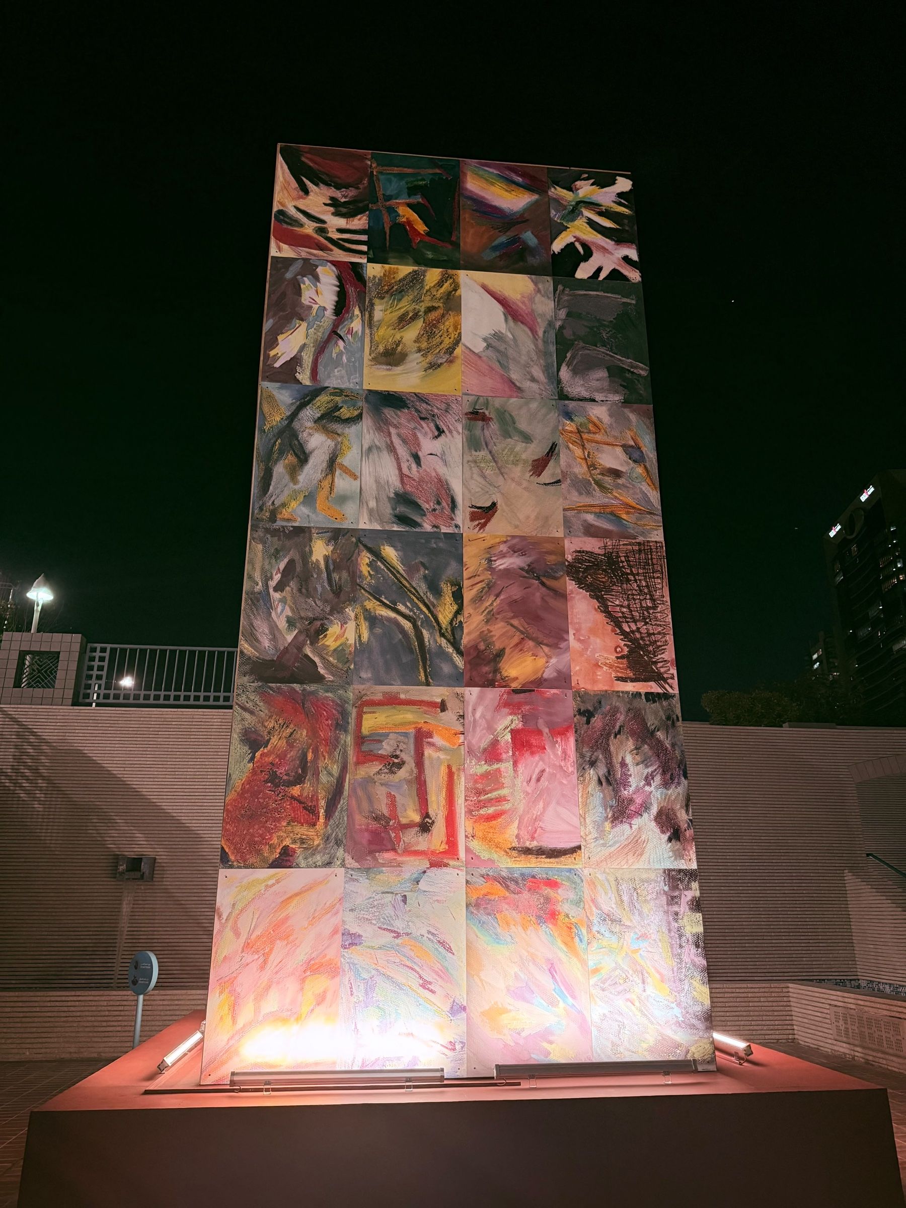 A tall stack of colourful abstract painted panels lit from below at night, standing on a plinth against a dark sky.