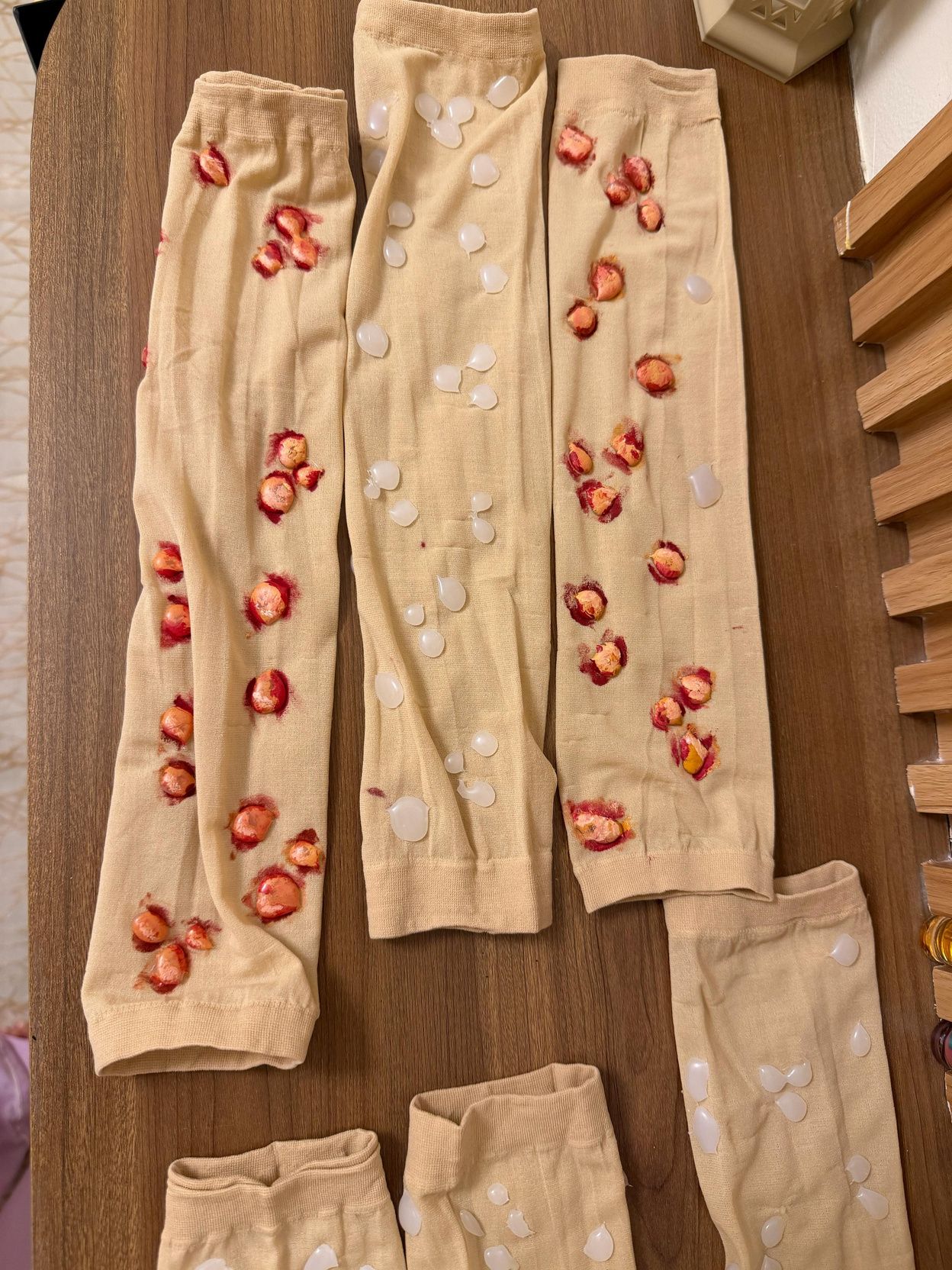 Skin-coloured arm sleeves laid on a wooden floor, covered in red and pale blister-like marks made with a glue gun.