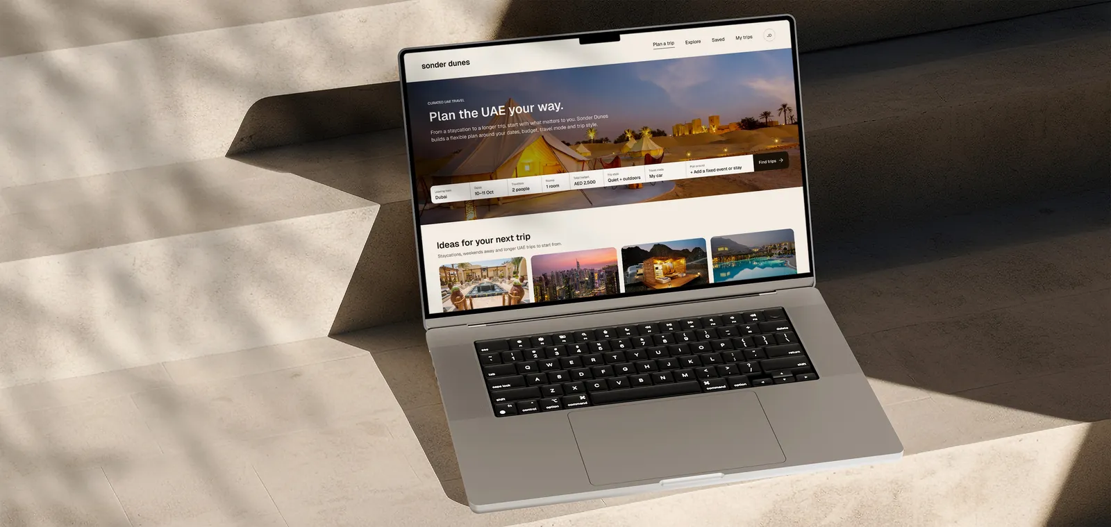 A MacBook Pro open on pale stone steps in dappled sunlight, showing the Sonder Dunes home screen: Plan the UAE your way, over a desert camp at dusk.