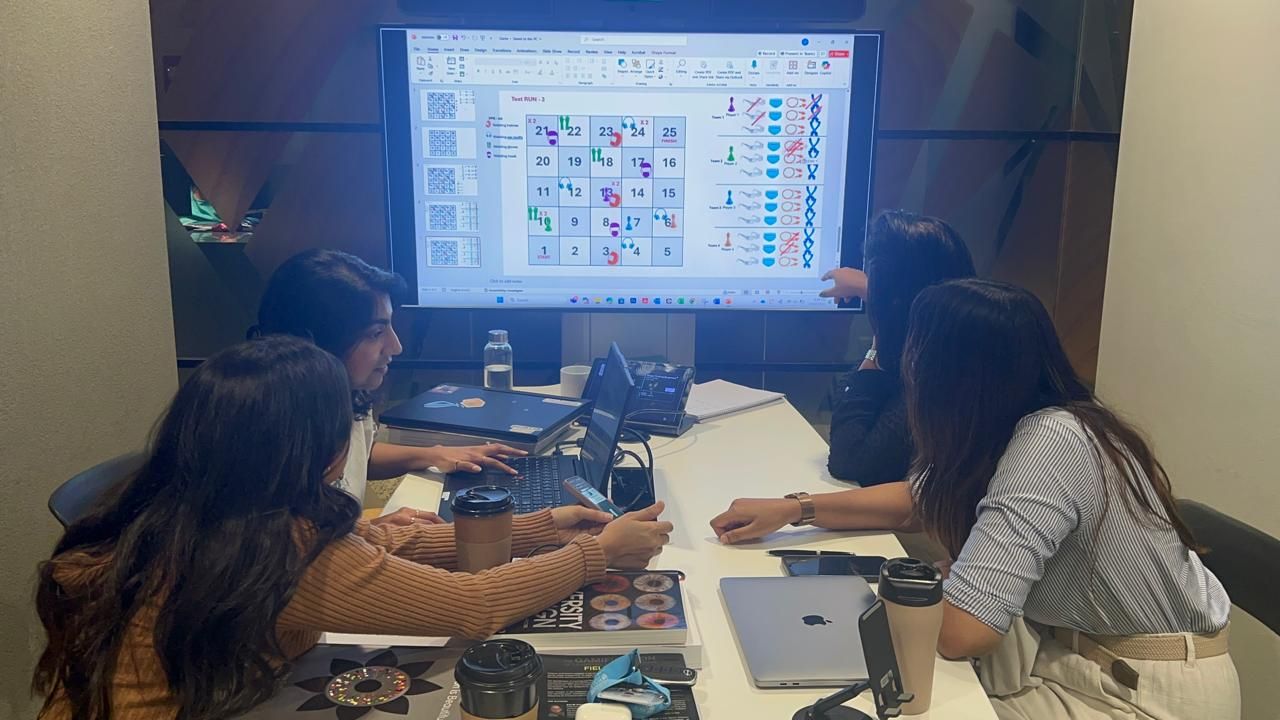 Team members playing the low-fidelity board around a table, with the digital board shown on a screen behind them.